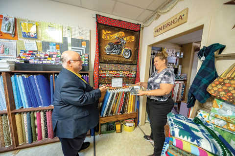 Man visiting quilt shop and talking with owner. Different fabrics are on the wall behind the two people.