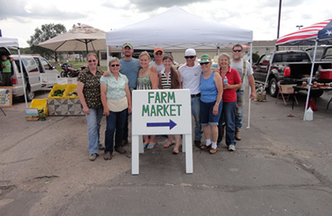 Group of people standing behind a Farm Market sign with vendor tents in the background.