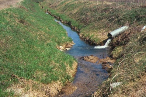 A metal pipe drains water into a stream in a gully on open farmland.