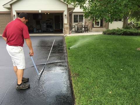 A person in a suburban yard measuring misdirected irrigation spray on a driveway.