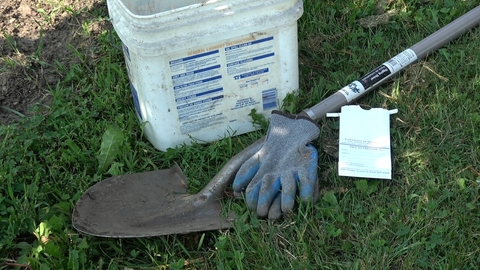 A long-handled metal shovel, a pair of blue gardening gloves, an old white bucket, and a paper bag from the UMN soil lab in the grass.