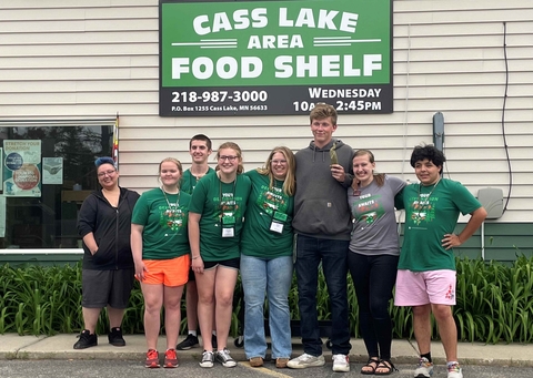 Eight youth stand close together in front of Cass Lake Area Food Shelf building and sign, several wearing 4-H t-shirts.