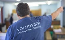 A person pointing with their back facing the camera wearing a shirt that reads volunteer well-water testing