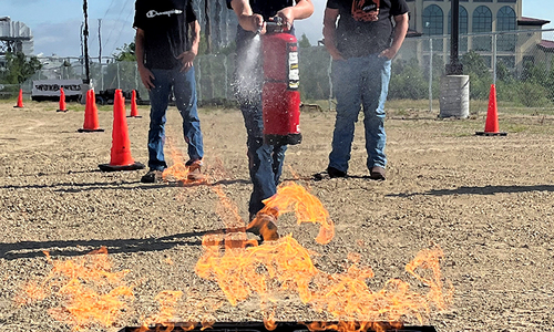 Three children practice using a fire extinguisher outdoors.