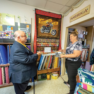 CJ Holl visiting in Kristi Feist in her Wells, Minn. quilt shop. Quilts and fabrics are on the wall behind them.