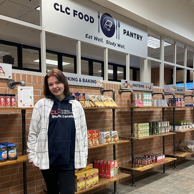 Student stands, smiling, in front of shelves of food. Her shirt says "CLC Student Senate"