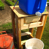 Handwashing stand built following these instructions, made of lumber with plywood top, and a square plastic water dispenser with a water catching bucket and trash can with foot operable lid