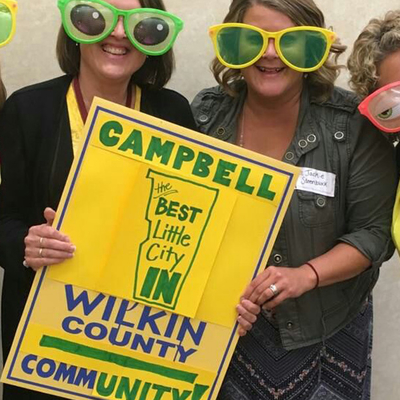 Four ladies wearing fun-oversized glasses holding a sign reading 'Cambell, the best little city in Wilkin County - CommUNITY'