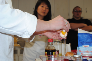 This image features a close up on the chef’s hands as she cracks open an egg. Other baking supplies is surrounding the table. Two women in the background are standing and watching the chef.